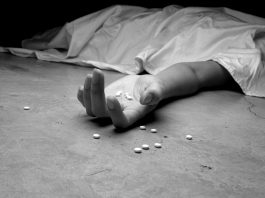 A black and white image of an addict, dead beneath a sheet, arm outstretched, pills in the palm of their lifeless hand and littering the floor near their hand.