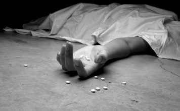 About this series – Addiction: A Rural Reality A black and white image of an addict, dead beneath a sheet, arm outstretched, pills in the palm of their lifeless hand and littering the floor near their hand.