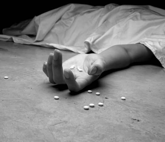 A black and white image of an addict, dead beneath a sheet, arm outstretched, pills in the palm of their lifeless hand and littering the floor near their hand.