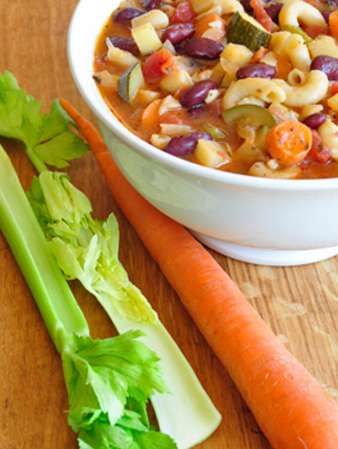 Minestrone Soup A bowl of minestrone soup with two pieces of celerey and one carrot on the table next to it.