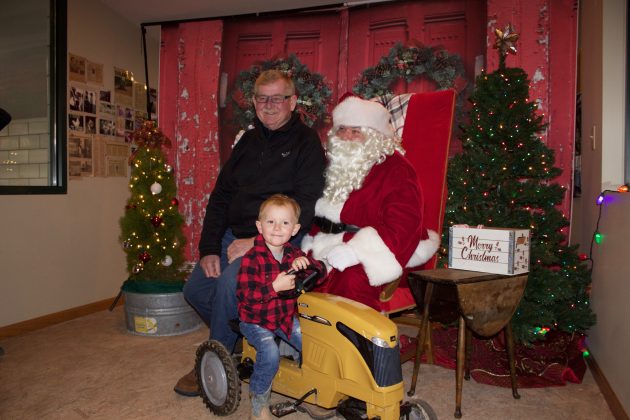 A grandfather sits on Santa's lap while his grandson sits on a yellow toy tractor in front of them.