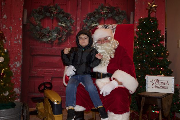 A boy with a black jacket and hood sits on Santa's lap for a picture.