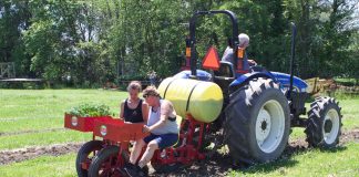 Two women sit in a vegetable planter while a man drives the tractor attached to it.