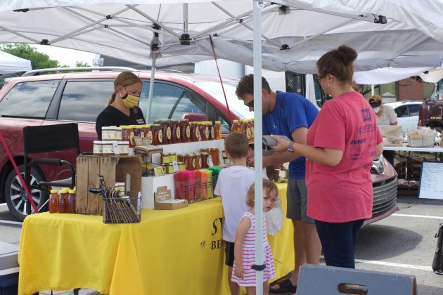 A woman serves customers from behind a stand at a farmers market.