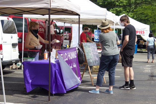 A woman serves customers from behind a stand at a farmers market.