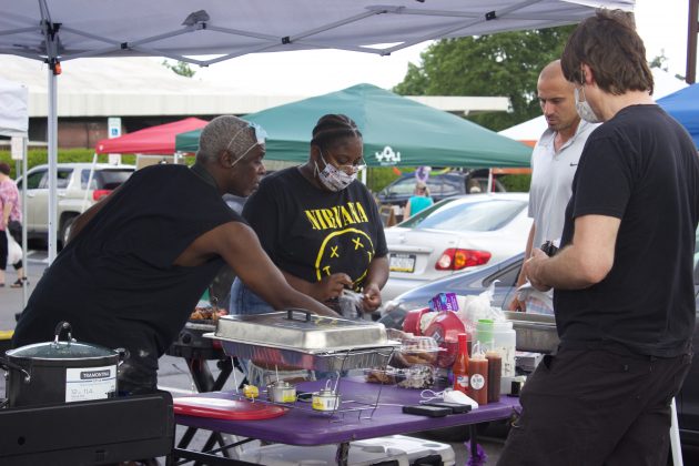 A man and woman serve customers from behind a stand at a farmers market.