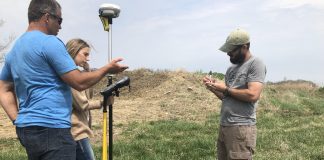 Three people stand in a field with GPS surveying equipment.