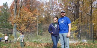 A woman and a man stand in front of a garden in the fall while a girl and a boy play in the background.