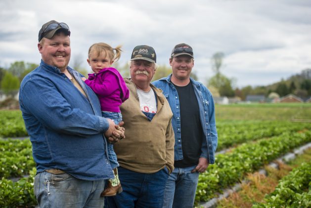 Three farmers stand on their farm, one of them holding his 2-year-old daughter.