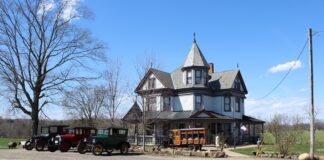 antique cars sit outside a victorian home. A home built hearse sits in front of the home with a casket in the back.