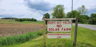 a sign reading roofs not fields no solar farm sits along a road with farm fields in the background