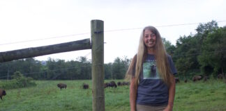 A woman stands in front of a fence, with bison on the other side.