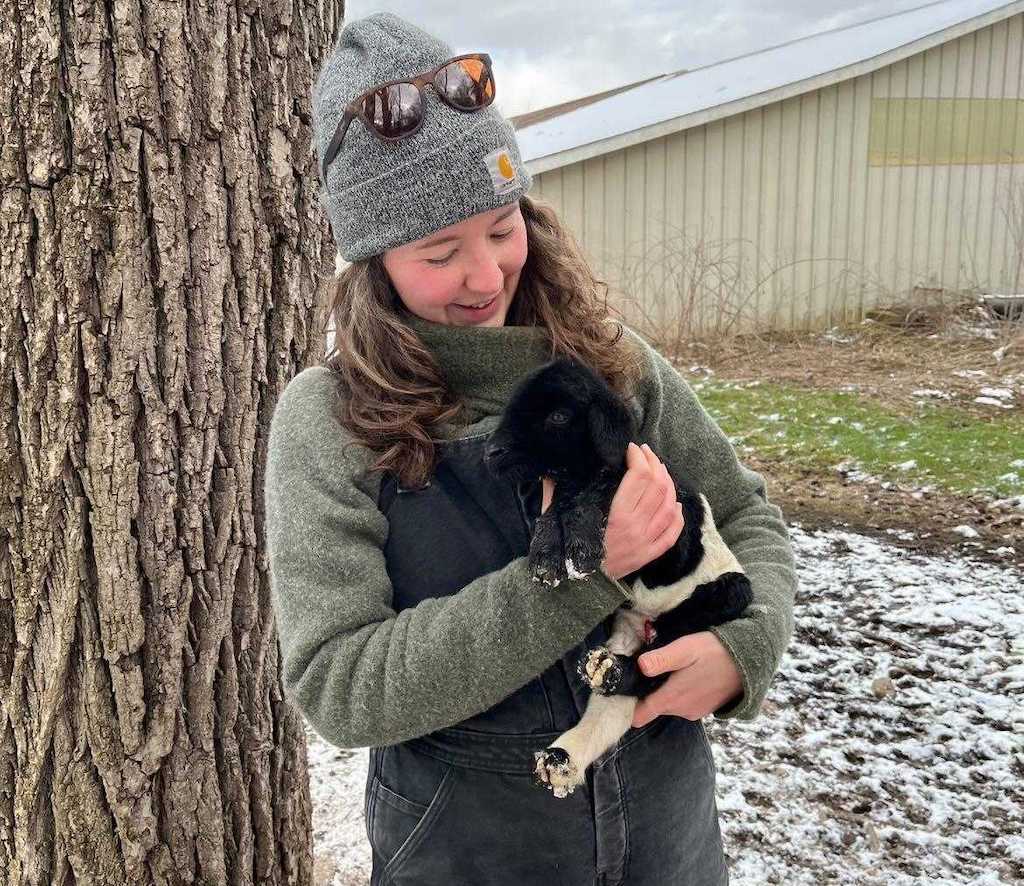 A woman in heavy overalls and a winter cap holds a small black and white lamb in her arms. The woman is smiling.