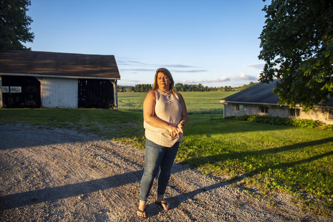 Farm family copes after suicide A woman wearing jeans and a light-colored top stands on a gravel driveway, framed in the background by a small open barn and a tree. Behind her are soybeans in a field.