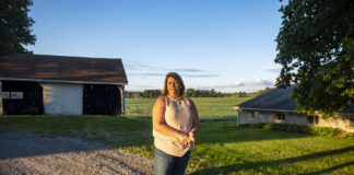 A woman wearing jeans and a light-colored top stands on a gravel driveway, framed in the background by a small open barn and a tree. Behind her are soybeans in a field.
