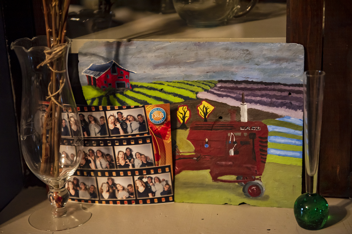 A string of photos sit on a shelf. Behind it is a painting of a tractor in a farm field.