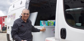 A man holds a cup of ice cream next to a van with freezers inside it and a cow pattern painted on the outside.