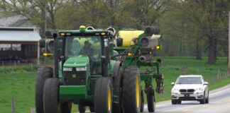 A tractor pulling a planter drives on a road with a car following closely behind it.