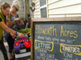 Two women stand next to table full of plants with sign in the foreground that says Ellsworth Acres