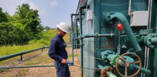 A worker holds a handheld gas detector up to a gas separator at a well pad