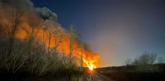 A flame reaching high into the sky can be seen at the end of a railroad tracks that go up the middle of the photo.